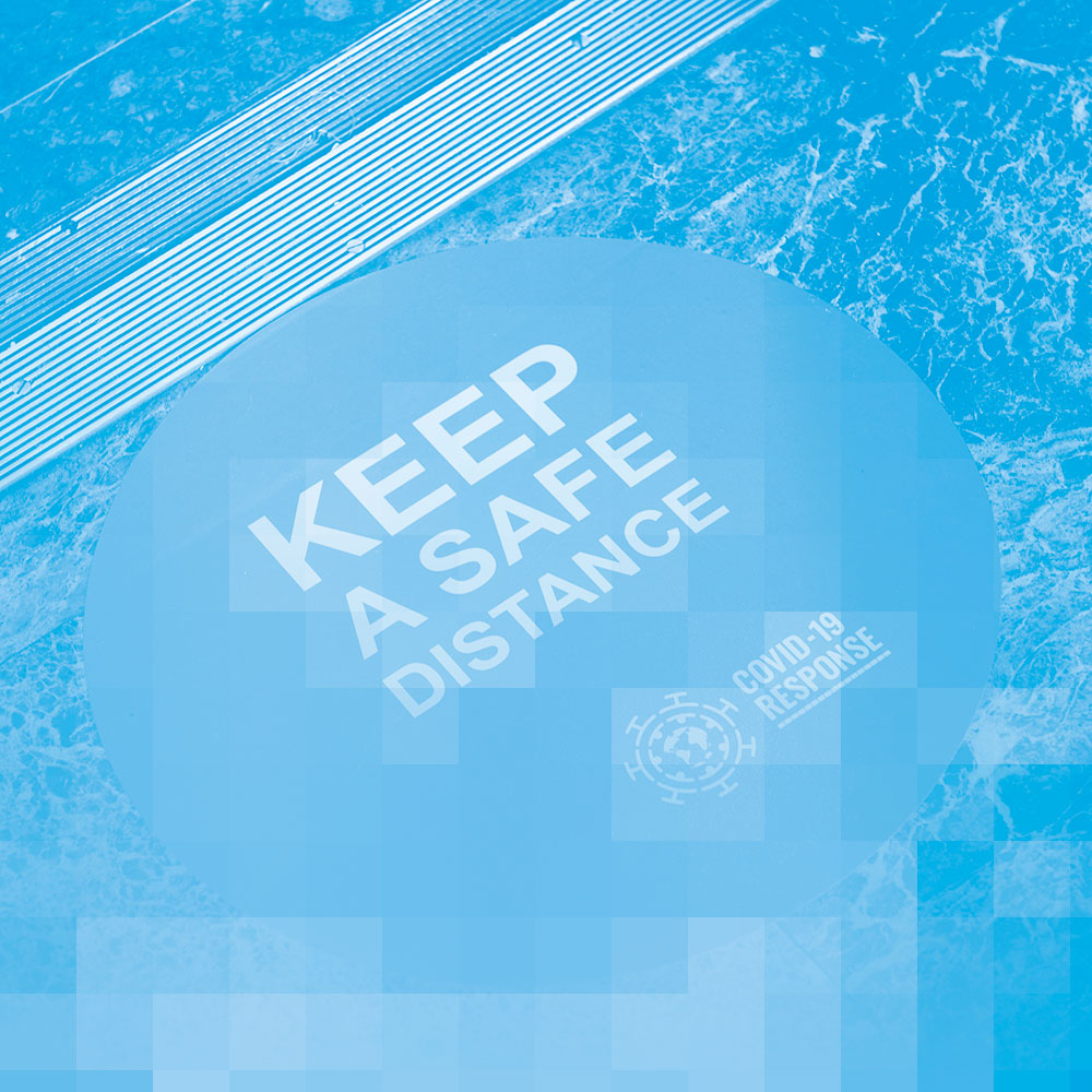 A photo, tinted in blue, of a large, round sticker on a floor. The sticker says “Keep a safe distance” and “COVID-19 response”.