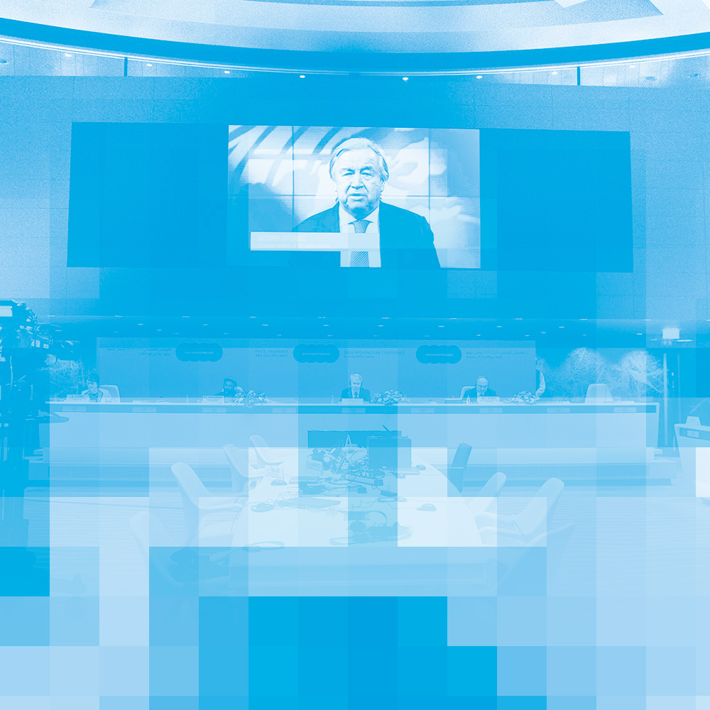 A photo, tinted in blue, of the podium of a conference room. People are seated apart at the podium. A screen above the podium is showing a video of the Secretary-General of the United Nations. On the left is a television camera directed at the podium.