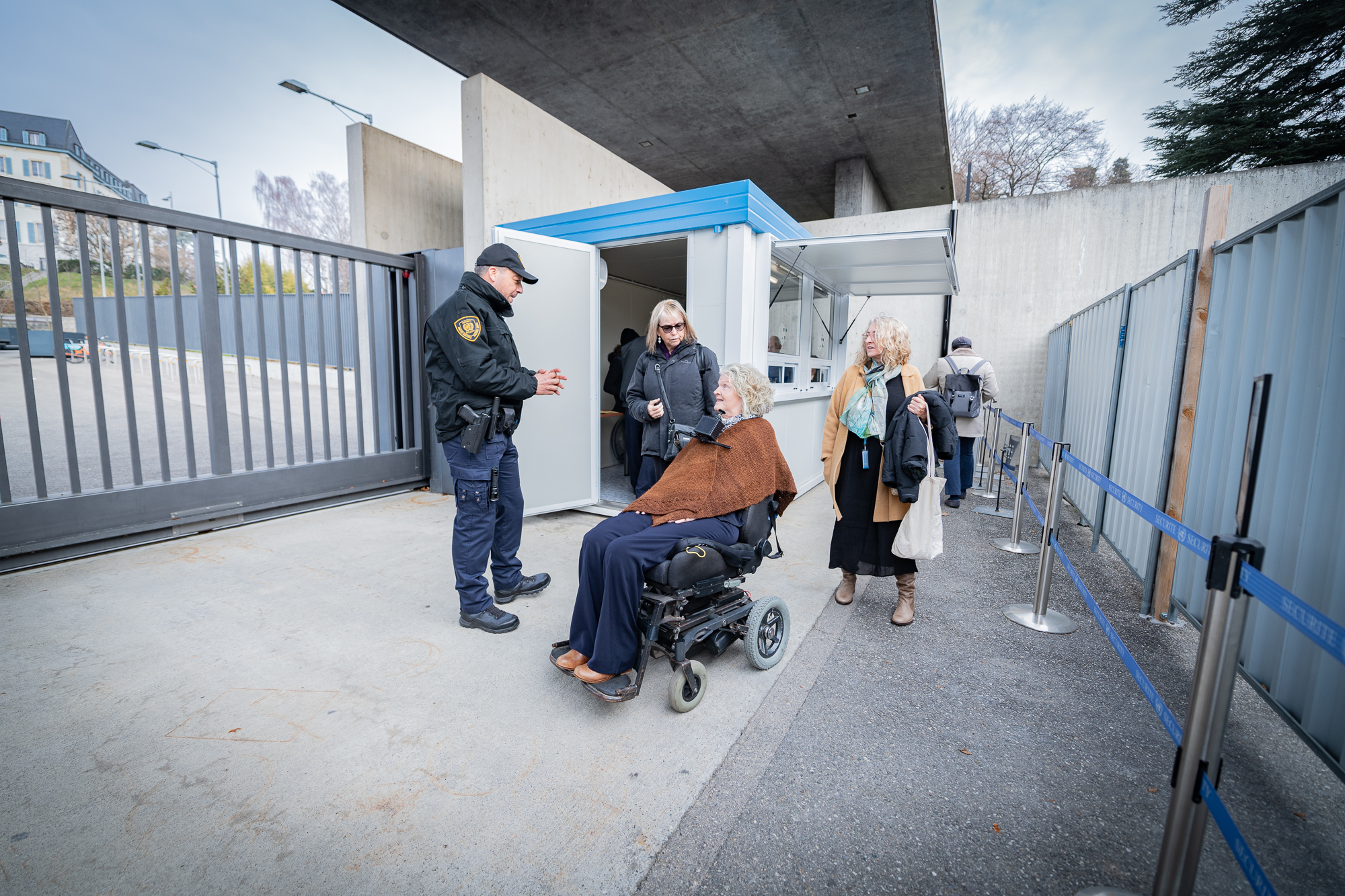 Next to a large grey gate, a Security and Safety Service officer speaks with a woman who is using a wheelchair. Two other women stand behind her.