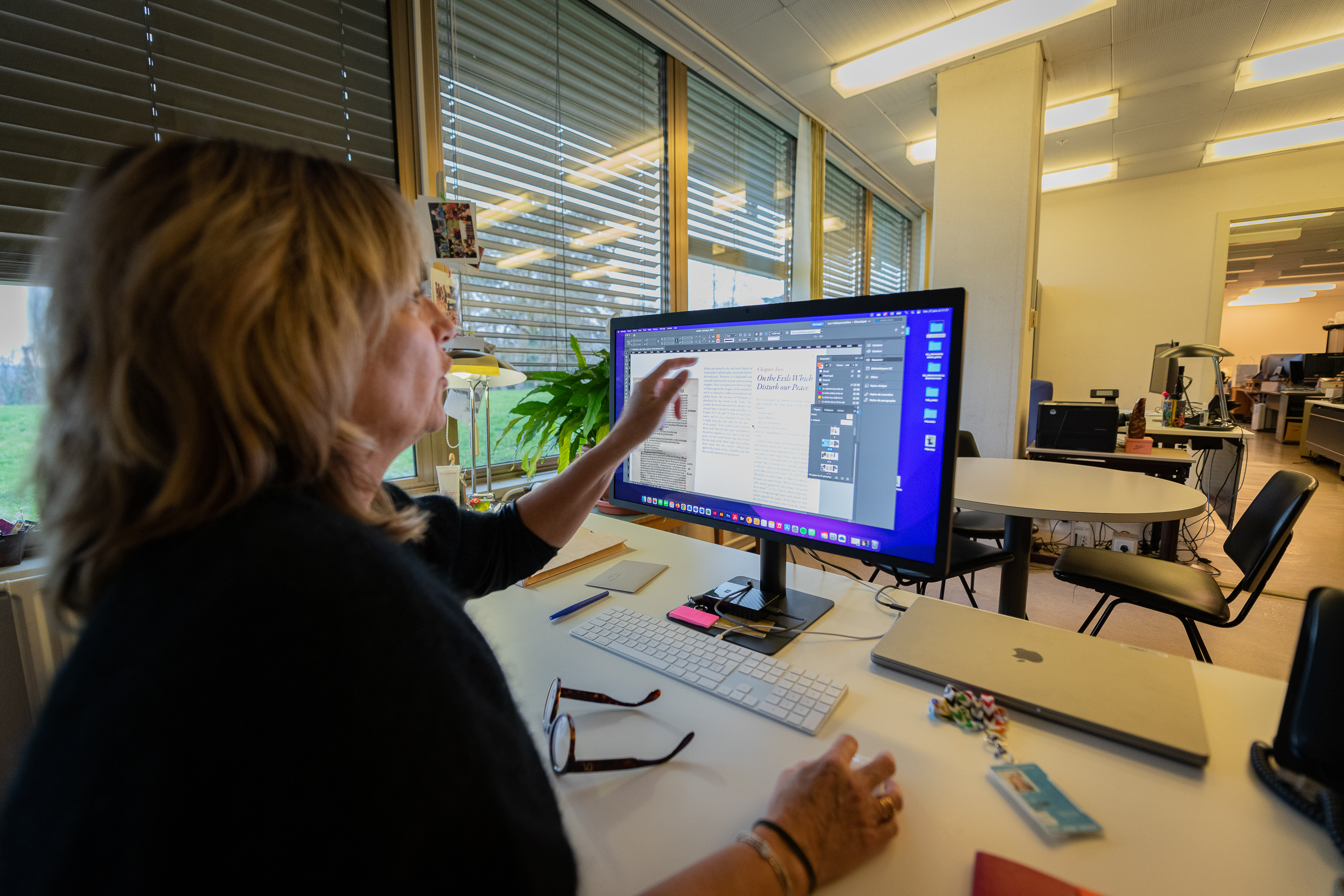 Une femme est assise à un bureau dans un grand espace de travail. Elle utilise un ordinateur et pointe l'écran du doigt.