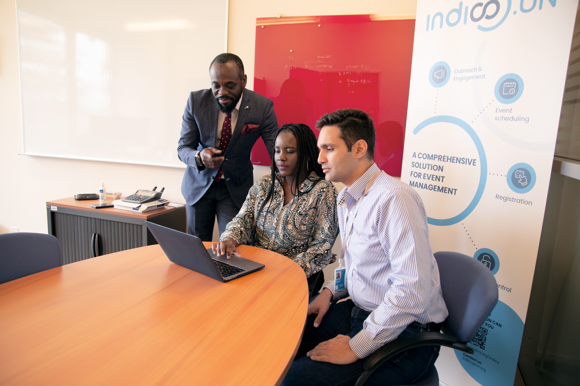 A woman sits at a wooden table using a laptop, while two men are on either side of her, watching. Behind them are a white board, a red glass whiteboard with writing and a poster with Indico branding.