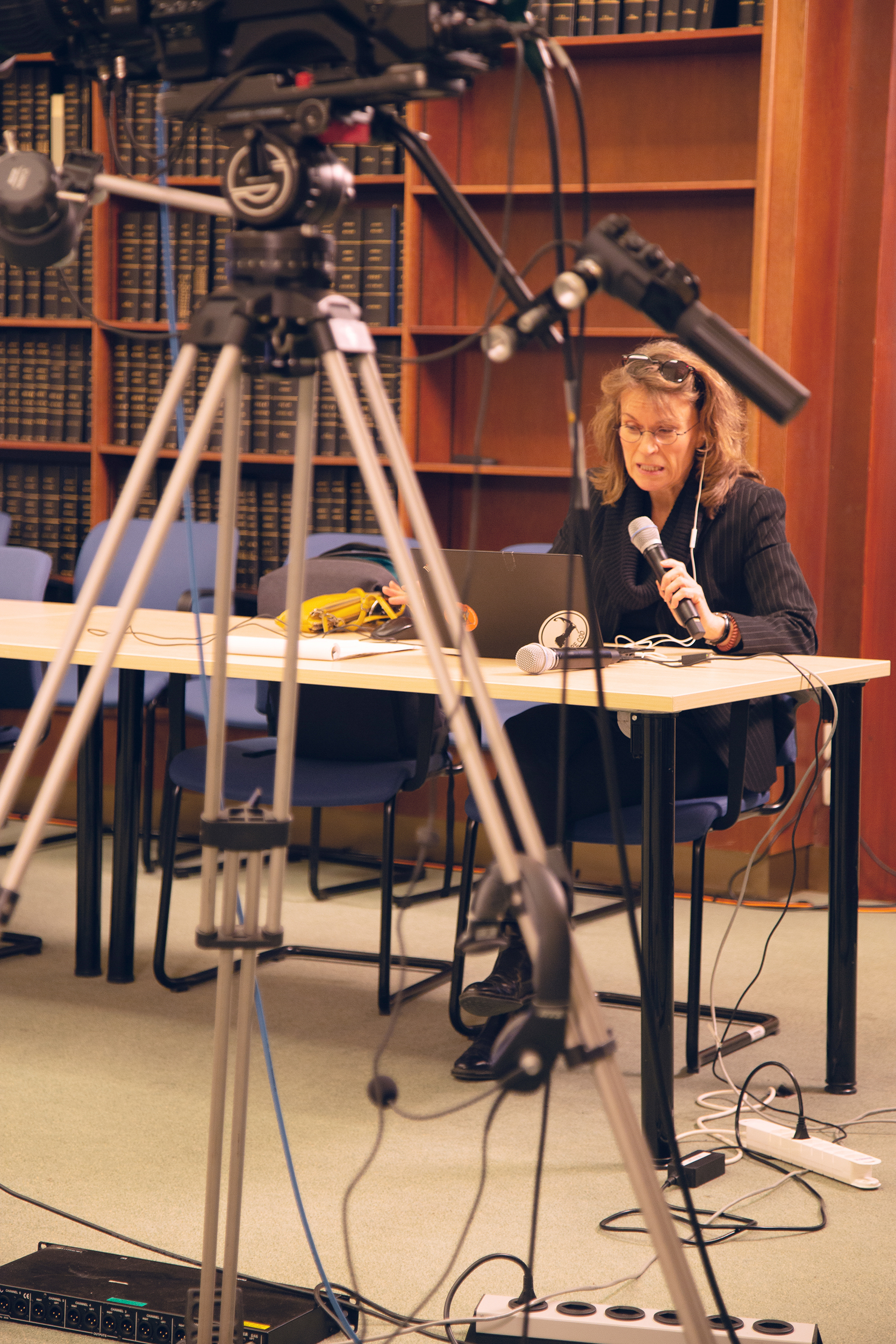 In the foreground of the photo is a tripod, surrounded by cords and other equipment. In the background, a woman is seated at a table and speaking into a handheld microphone. In front of her, on the table, is another microphone. Behind her are chairs and wooden shelves with books. 
