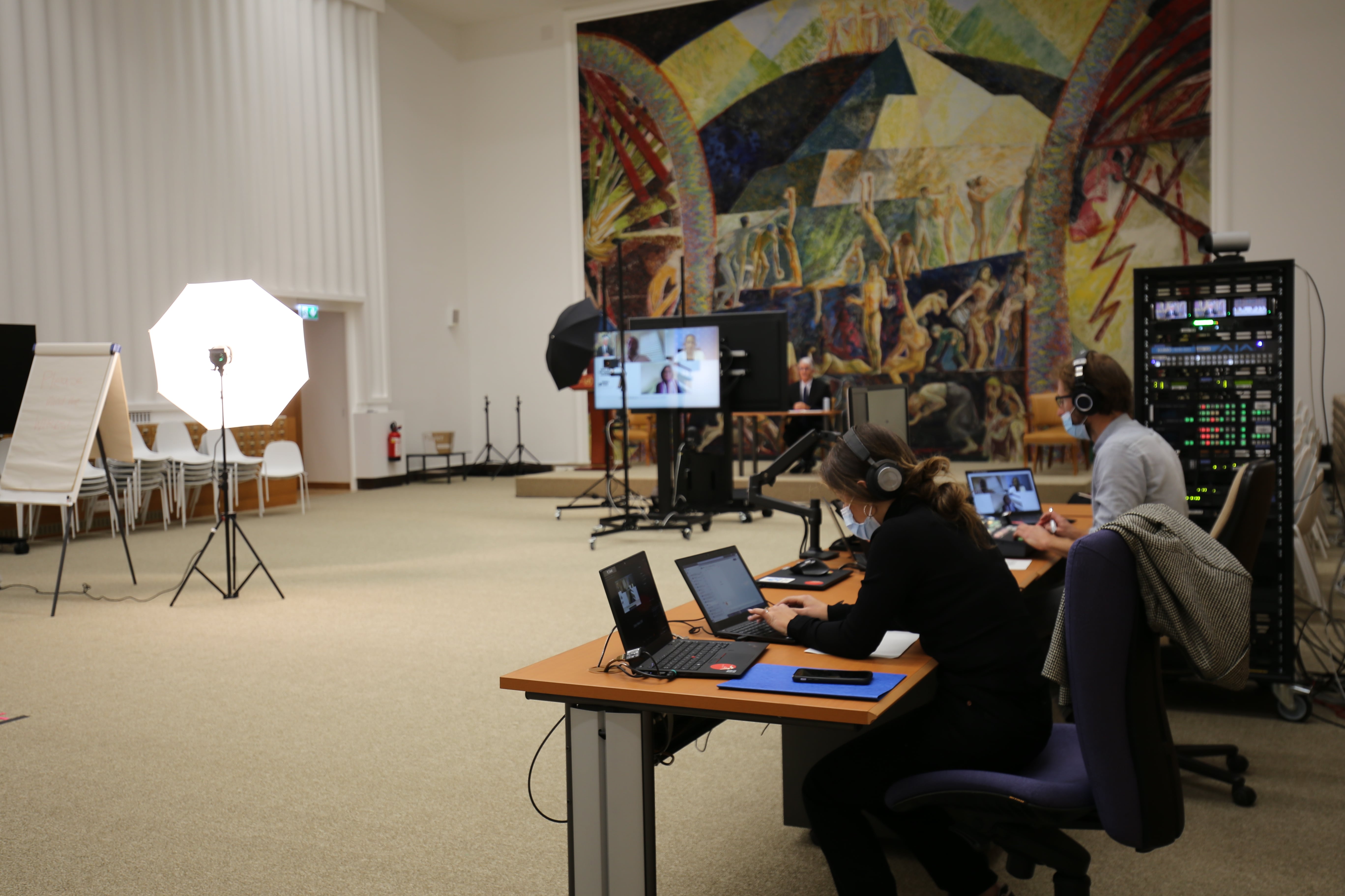 A photo of a large room with a mural at the end of it. On the right, two people are seated at a long desk. They are working at laptops in front of them and wearing headphones and facemasks. More equipment is next to them. On the left there is a flip chart and lighting equipment. At the back of the room, in front of the mural, is a large television screen and more lighting equipment.  