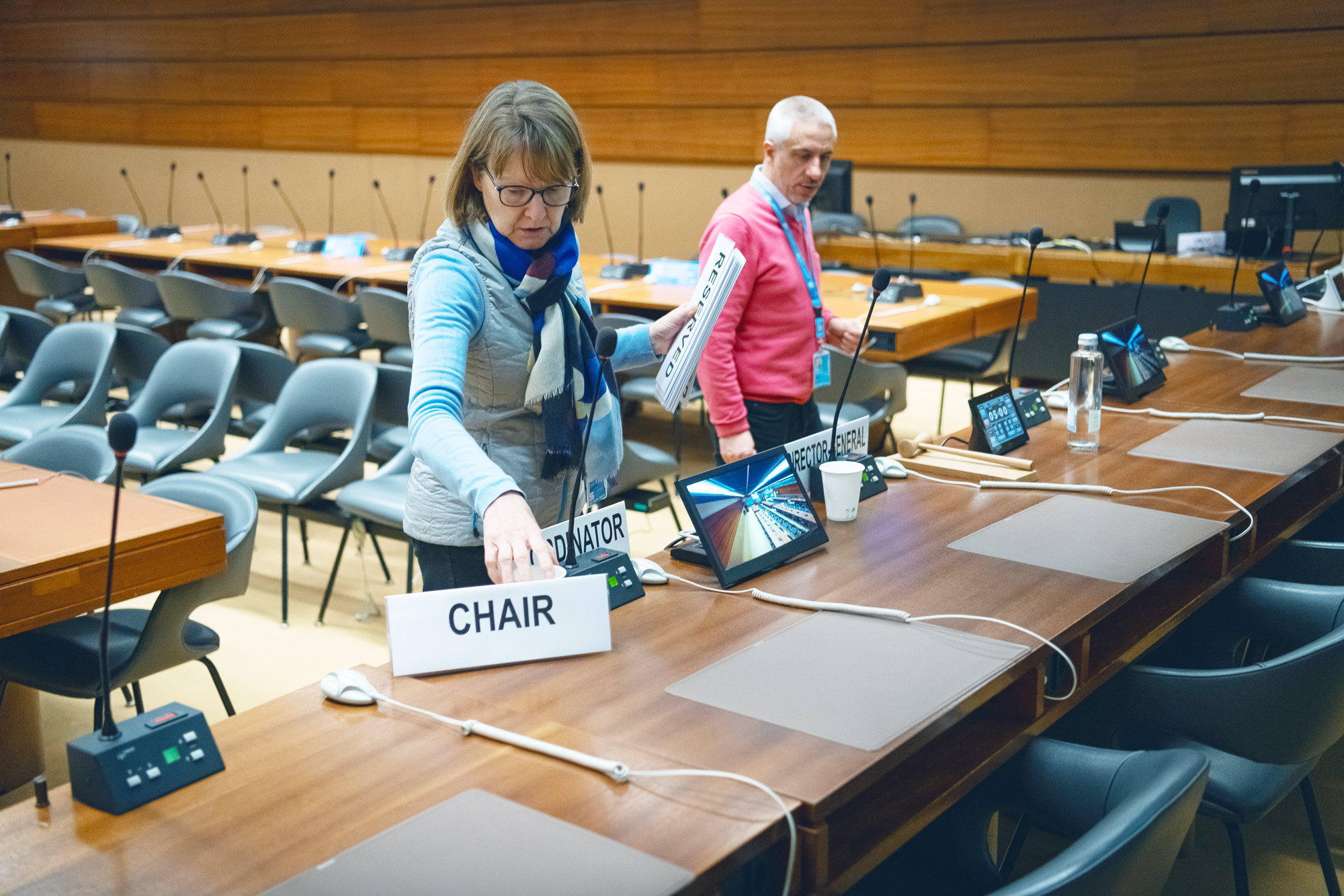 A woman places a nameplate saying “chair” on a wooden table in a conference room near a nameplate that says “coordinator”. In her other hand is another nameplate that says “reserved”. A man stands near her. In the background there are two long wooden tables with microphones and rows of chairs. 