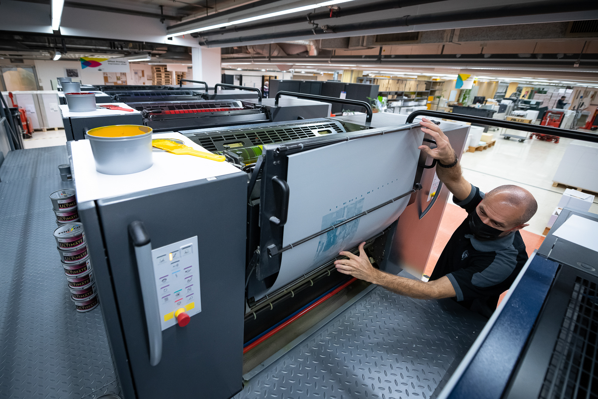 Photo prise en plongée d'un homme utilisant une imposante machine à imprimer dans un espace ouvert. Il regarde un grand document sur la machine à imprimer.
