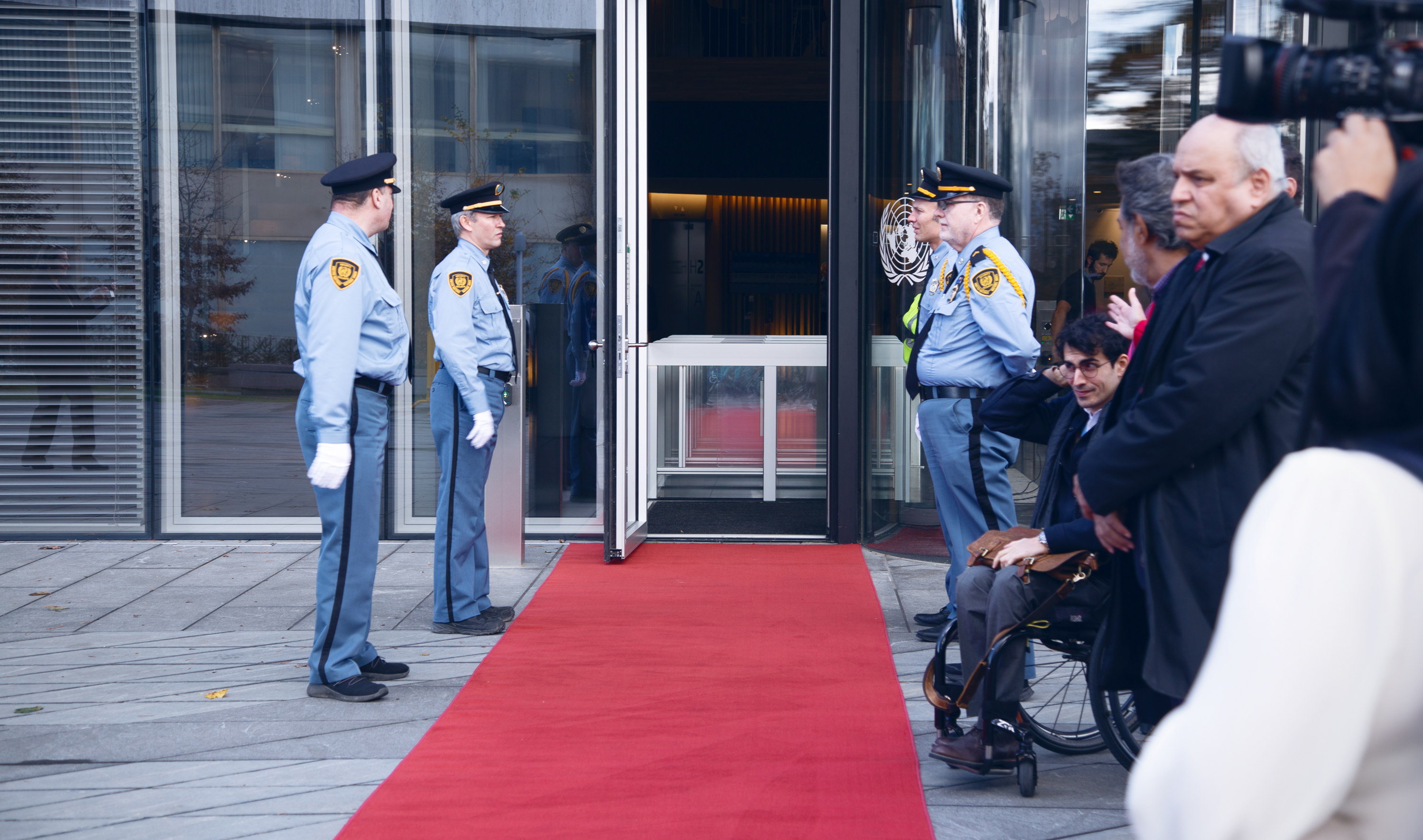 A red carpet is laid out in front of an open door to a glass building. Four Security and Safety Service officers stand on either side of the red carpet, two on either side. On the right, there is a small crowd of people and a television camera. 