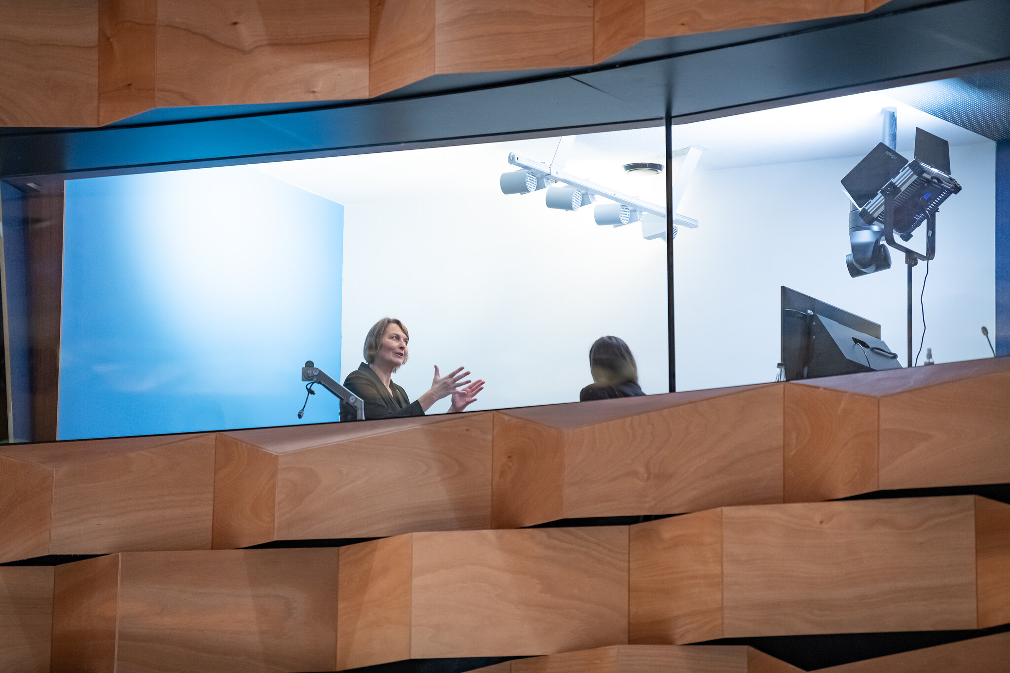 A photo taken from below, looking up to a windowed room. Two women are facing each other inside the room. The woman on the left stands in front a blue wall, gesturing. On the right is television screen and lighting equipment. 