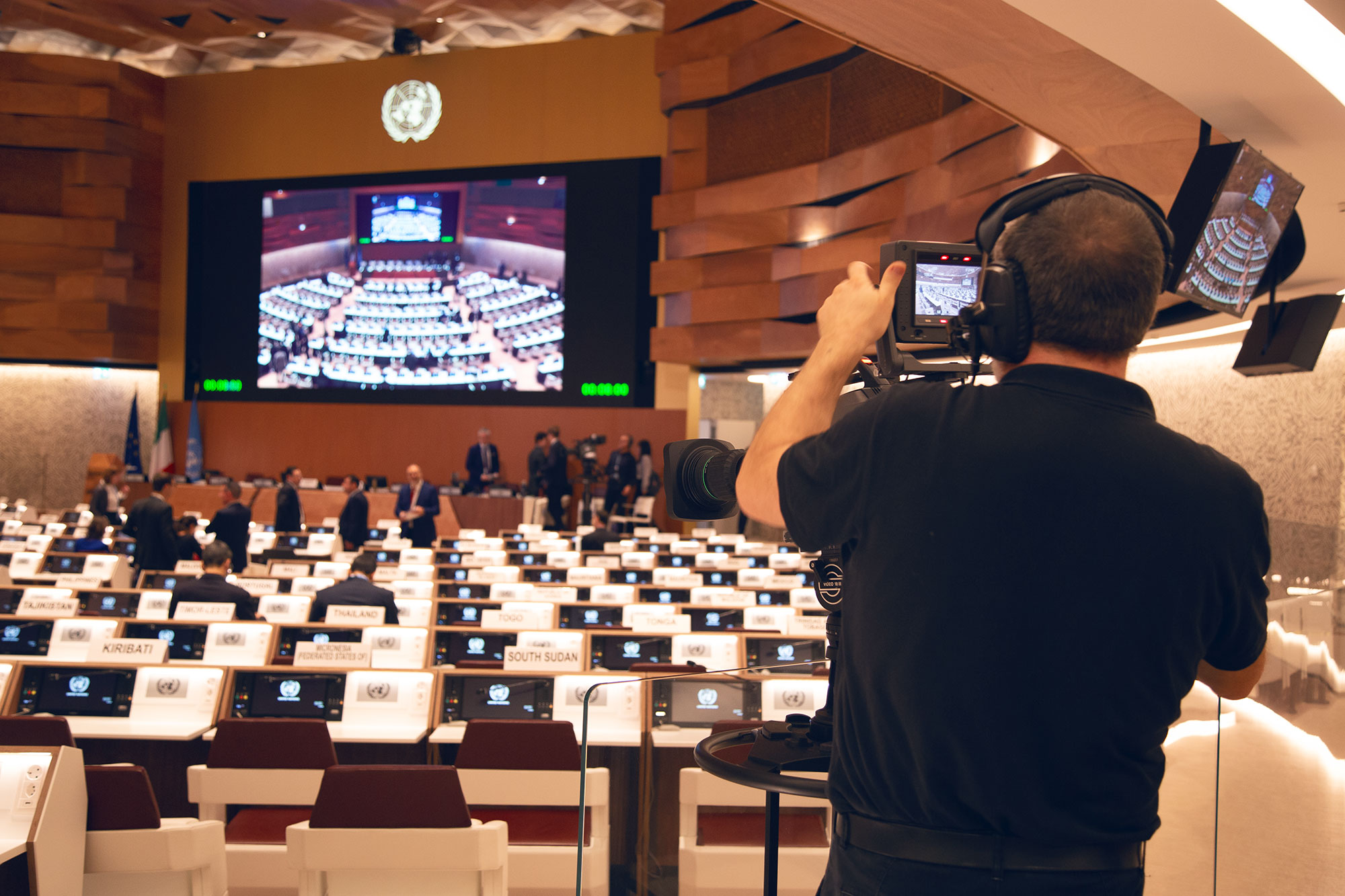 A photo from the back of a large conference room. At the front of the room, above the podium, is a large screen showing a shot of the conference room. People are mingling near the podium and first rows of desks and chairs. In the foreground of the photo, facing away, is a man operating a video camera and wearing headphones. 
