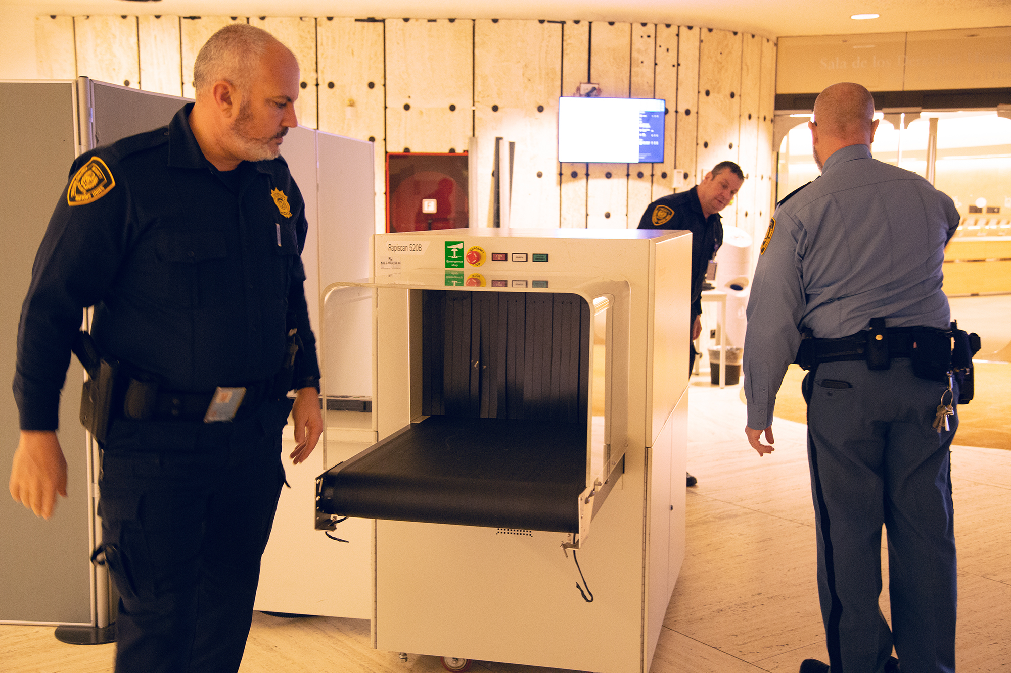 Three Security and Safety Service officers surround an x-ray machine. In the background there is an entrance to a conference room and a television screen mounted on a wall.
