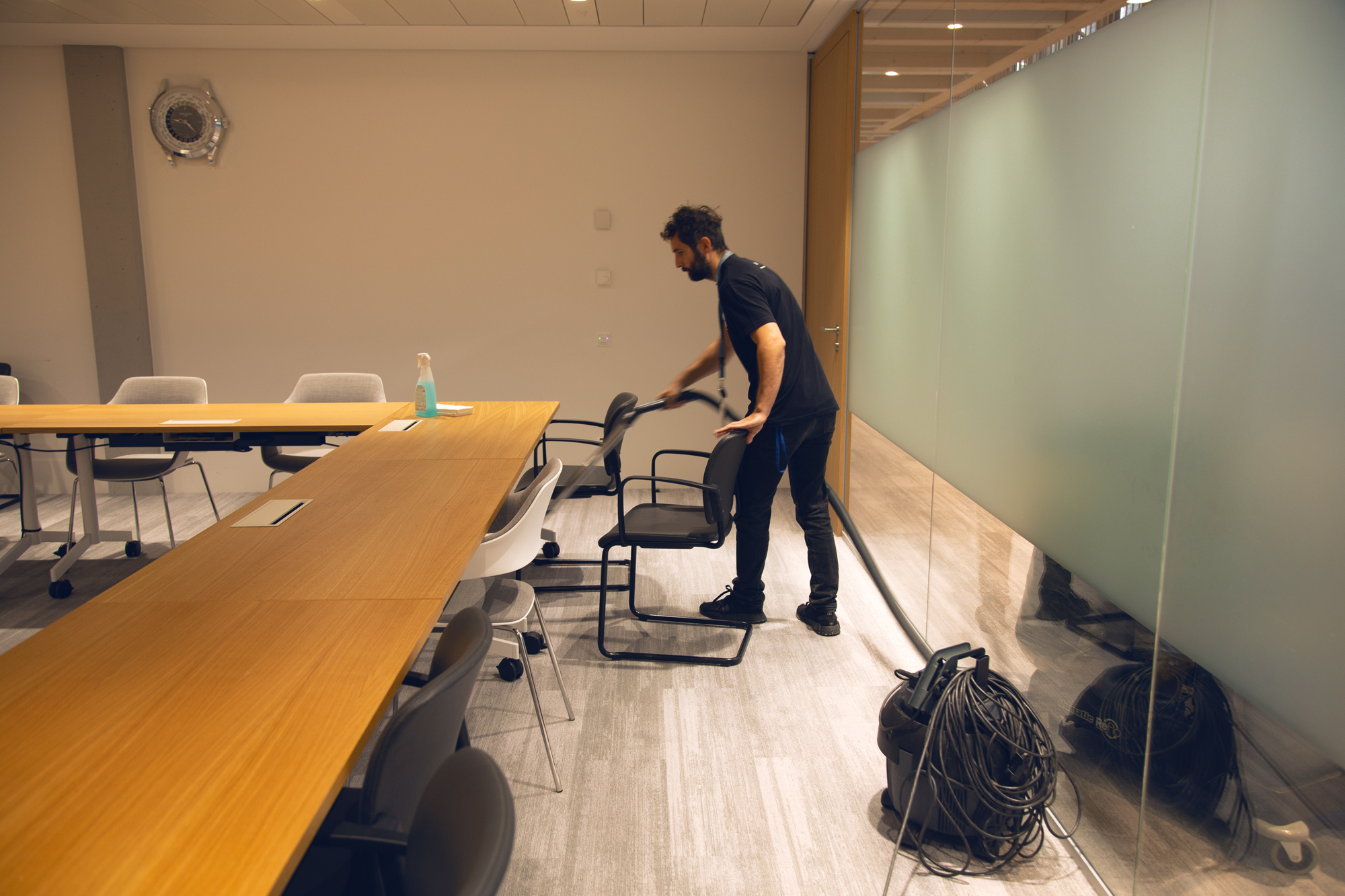 A man vacuums a meeting room. He is moving one of the chairs that surrounds a large wooden table. On the right is a partially-frosted glass wall. 