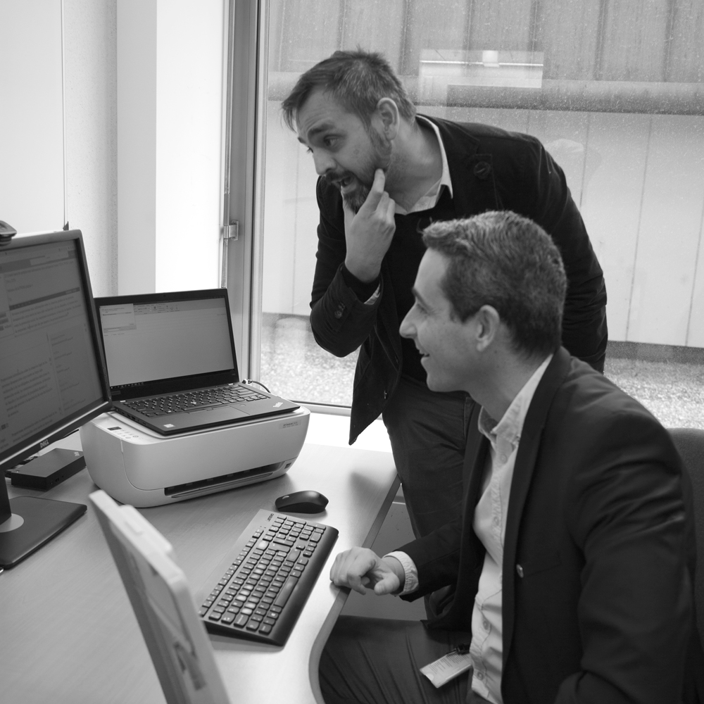 Two men work at a desk; one man is sitting while the other is standing. They are working at a computer and looking at several monitors. There is also a laptop on the desk.
