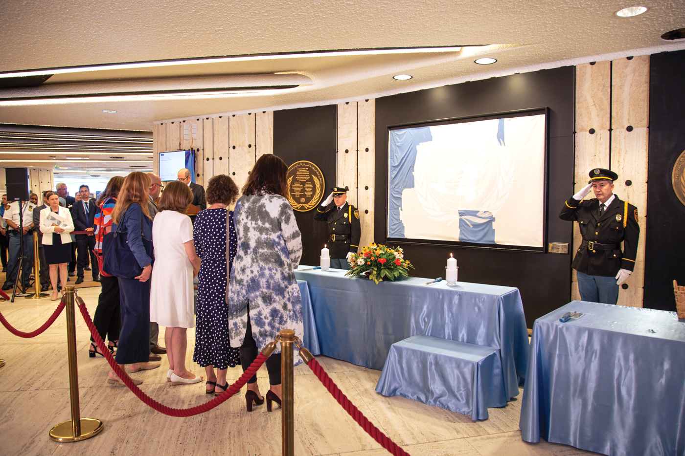 A group of five women is standing in front of a table covered by a light blue cloth. They are bowing their heads. A flower arrangement and two lit candles are placed on the table. A frame with the remainder of a United Nations flag is hanging on the wall behind the table. On each side of the table are two security officers. 