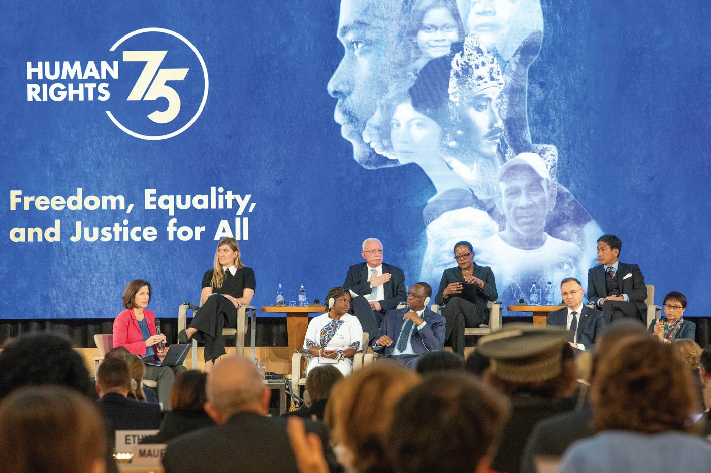 Photo of a panel discussion taking place in a large conference room. A woman is moderating a discussion among eight panelists. An audience is visible at the forefront of the photo. In the background is a large dark blue banner with white portraits of people, the ‘Human Rights 75’ logo and the words ‘freedom, equality and justice for all’ written in white. 