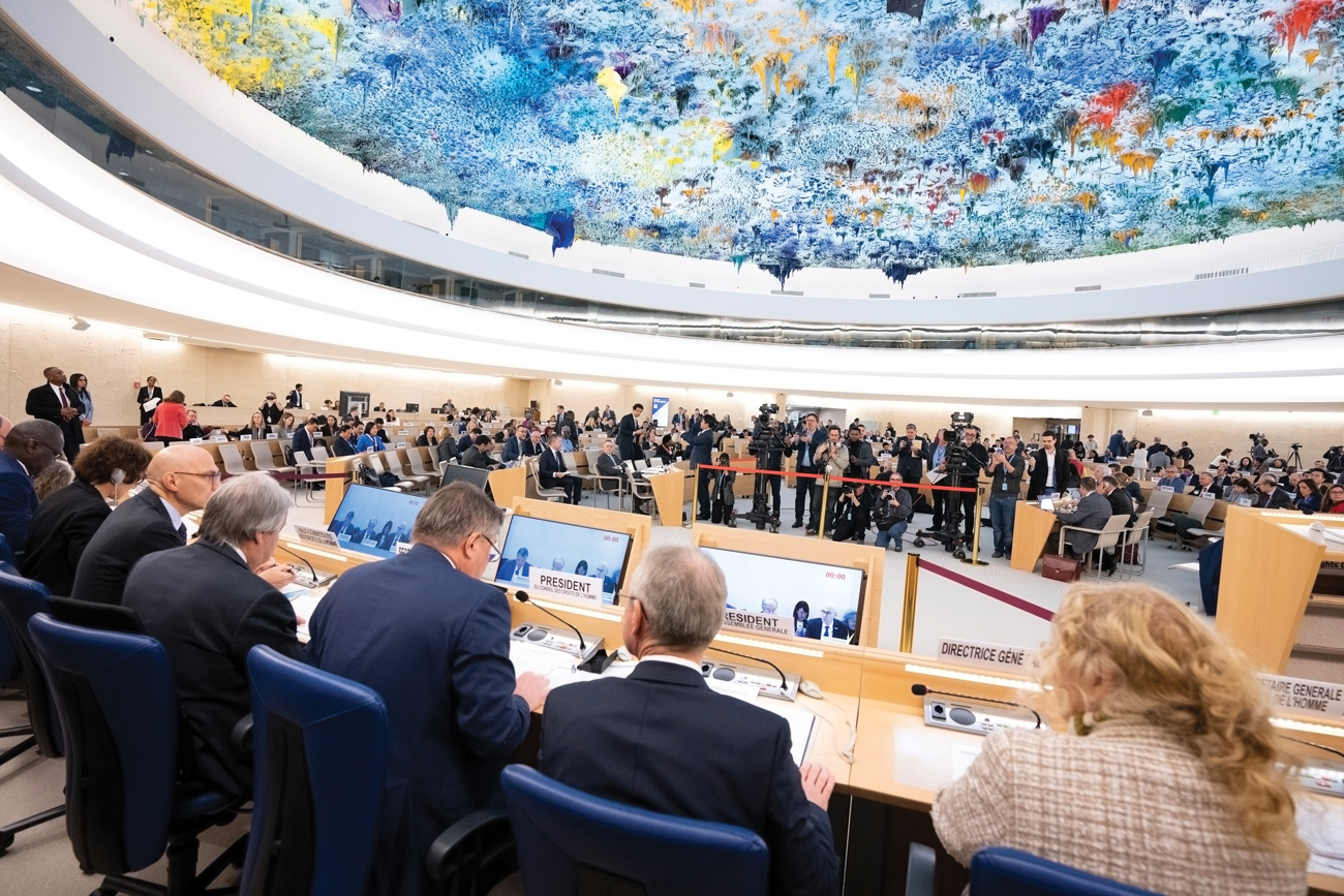 Back view from the Director-General of the UN Office at Geneva, Tatiana Valovaya, the President of the UN General Assembly, Csaba Kőrösi, the President of the Human Rights Council, Václav Bálek, the Secretary-General of the United Nations, António Guterres, and the High Commissioner for Human Rights, Volker Türk sitting at a podium in a conference room. Journalists are facing them and taking photos of them. The ceiling of the room depicts a modern and vibrant artwork. 