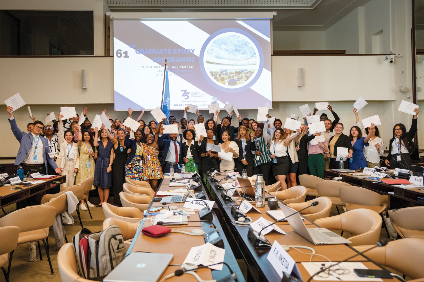 A diverse group of students is standing together in front of a wall where a screen is placed. All of them are holding their diplomas in their hands.  In front of them are desks and chairs where laptops and papers are placed. 