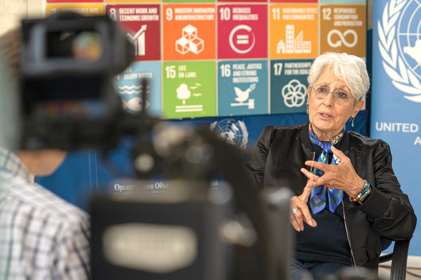 Singer and activist Joan Baez is sitting in a chair giving an interview. At the forefront, the camera is visible but blurred. We see part of the journalist on the left side. Behind Ms. Baez is a wall with square images representing the Sustainable Development Goals. 