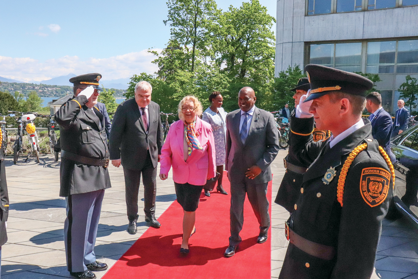 The photo shows the Director-General of the UN Office at Geneva wearing a pink jacket and a colorful scarf and welcoming the President of Botswana, Mokgweetsi Eric Keabetswe Masisi. Both of them are walking on a red carpet. The honor guard is standing on each side of the carpet. 