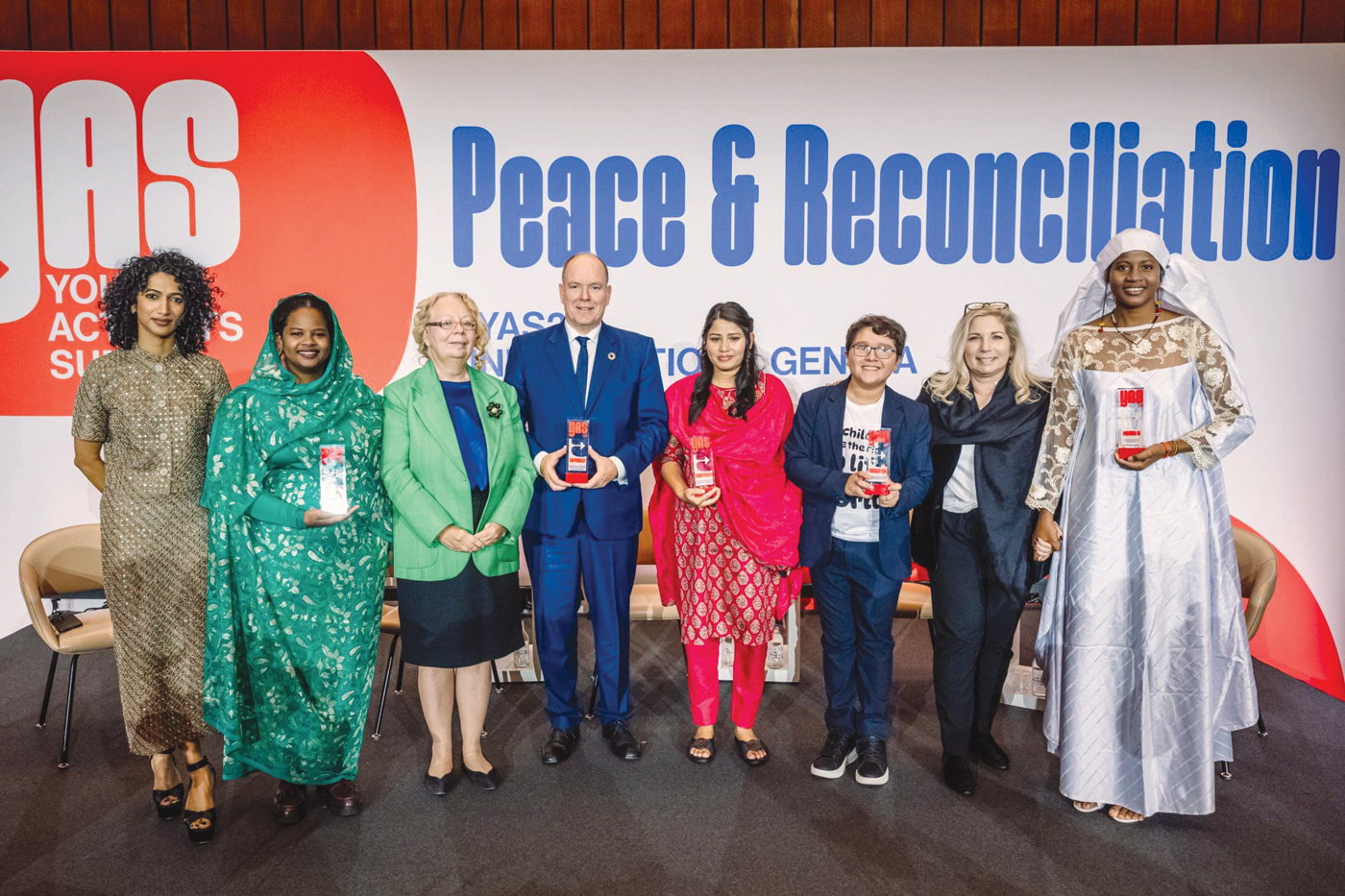 Un groupe de huit personnes se tient devant un mur où se trouve une grande banderole portant l'inscription « Young Activists Summit - Peace & reconciliation » (Sommet des jeunes activistes - Paix et réconciliation). Six femmes et deux hommes font partie du groupe, dont le prince Albert II de Monaco et la Directrice générale de l'Office des Nations Unies à Genève, Tatiana Valovaya. Le Prince et trois autres membres du groupe tiennent un prix.