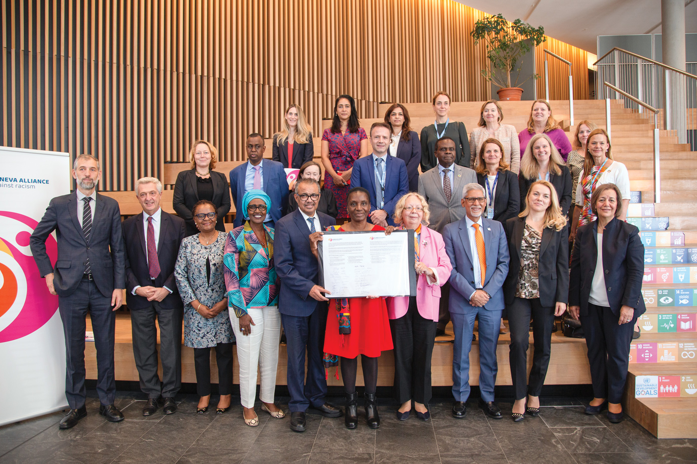 Un groupe diversifié composé d'hommes et de femmes se tient devant de grands escaliers en bois. Le groupe se tient sur trois rangs différents. Au premier rang, trois personnes tiennent un grand document.