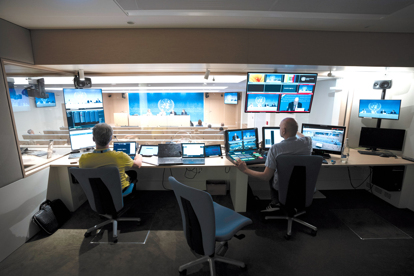 The photo shows two technicians running the audiovisual equipment in a technical room located above a press room. The technical room is filled with monitors and two control panels. The below conference room shows four persons sitting at a podium, speaking to an audience. 