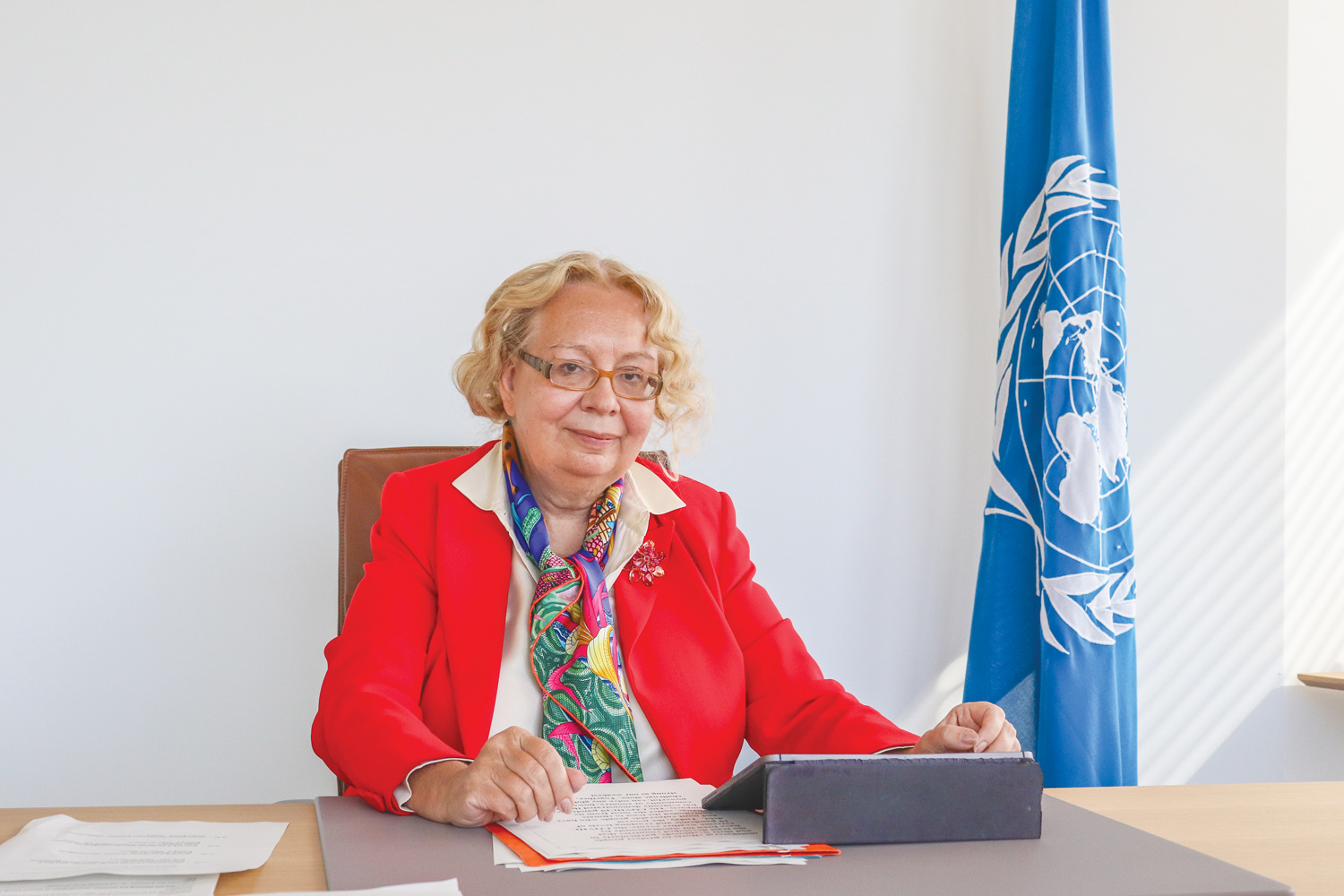 Photo of a blond woman wearing a red jacket sitting at her desk. She is looking at the camera. Behind her, on the right, is a United Nations flag. An iPad and some papers are placed on the desk. 