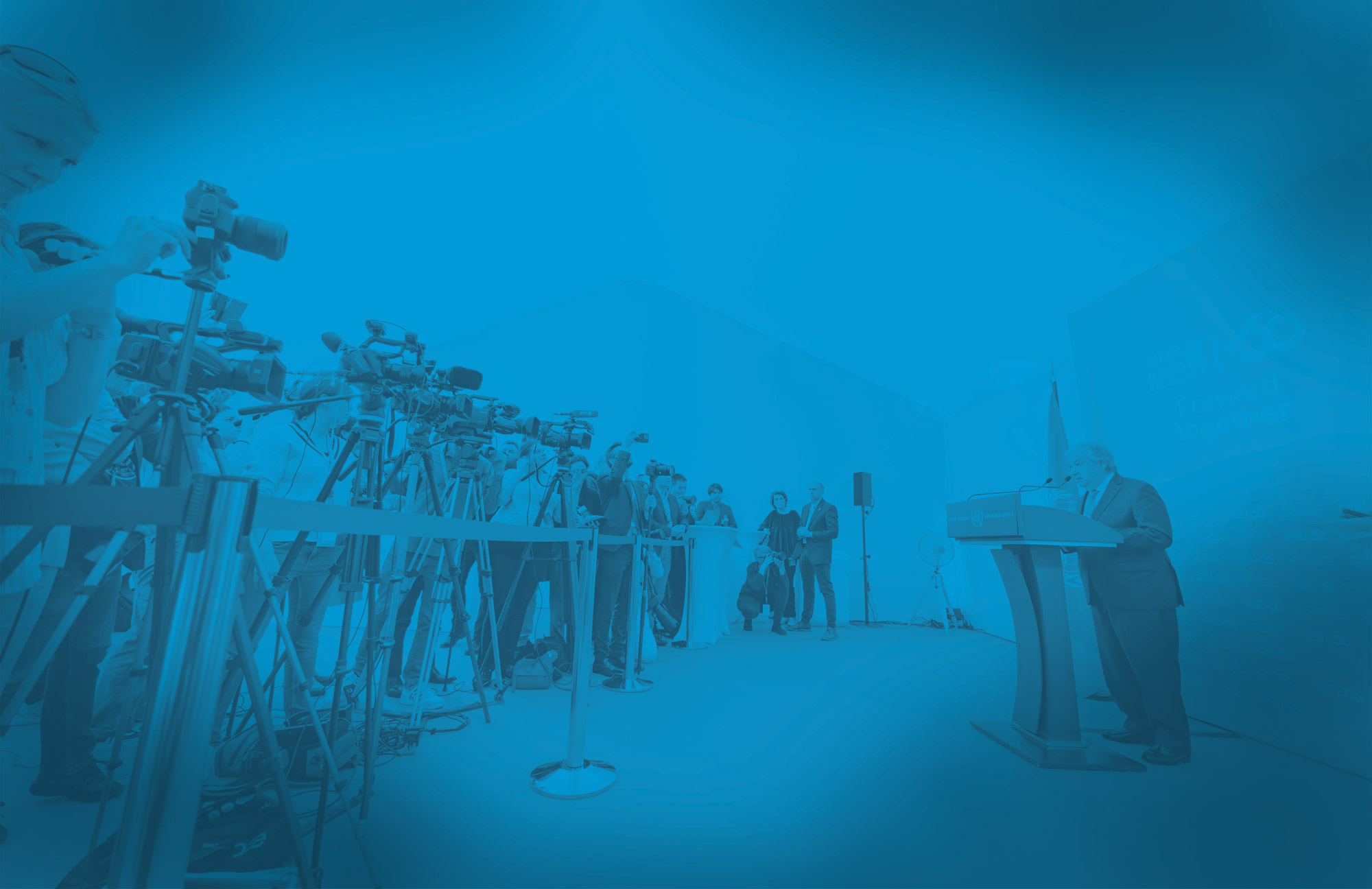 A man is standing behind a rostrum and speaking to a pool of journalists. A UN flag is displayed by his side.