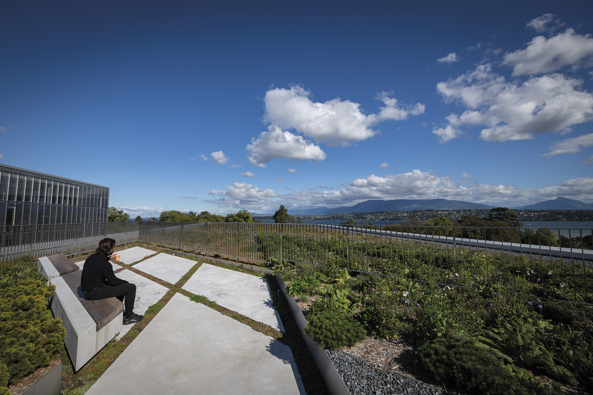  An employee sitting on the bench surrounded by low vegetation and looking at the view of Geneva, Geneva Lake, and mountains. 