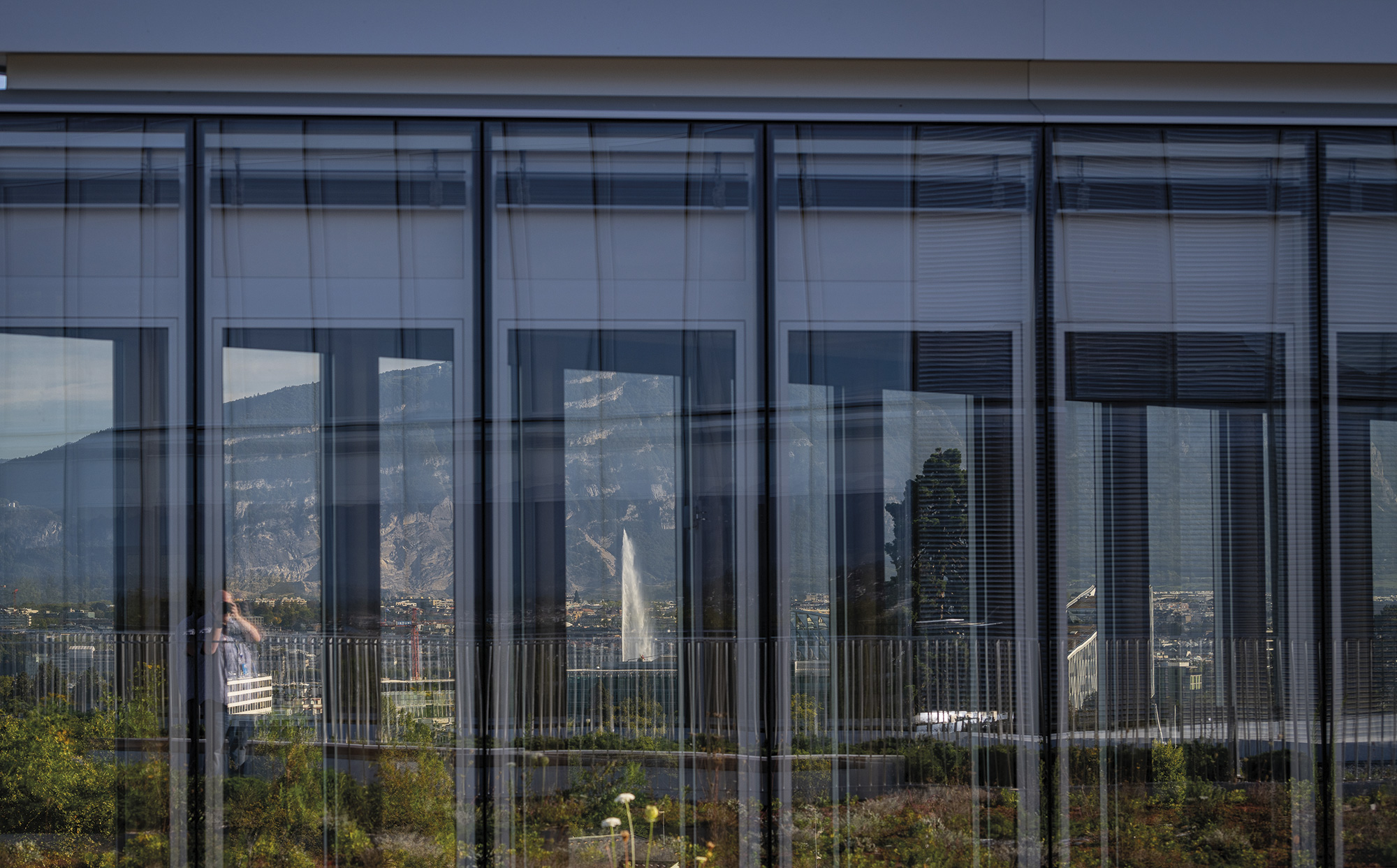 Windows on a new building reflecting mountains, Geneva town, and the large lake fountain.