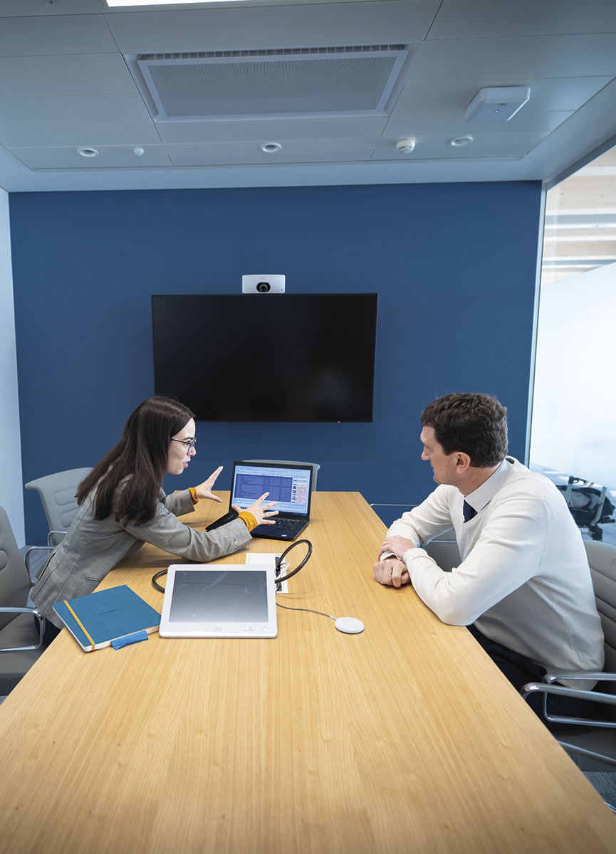 Employees sitting in a conference room at the desk with an iPad and a notebook and discussing the charts on the laptop which is placed between them..