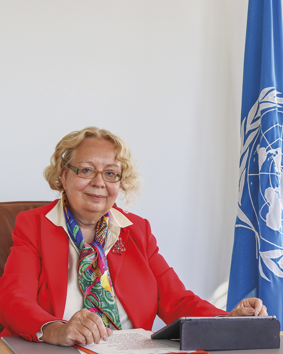 Director-General of the United Nations Office at Geneva, Tatiana Valovaya, sitting in the office with an iPad and papers in front of her. The United Nations flag in the background.